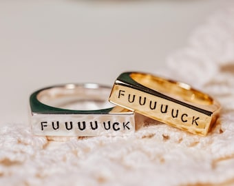 May include: Two silver and gold rings with the word "FUUUUUCK" engraved on them. The rings are stacked on top of each other and are sitting on a white crocheted surface.