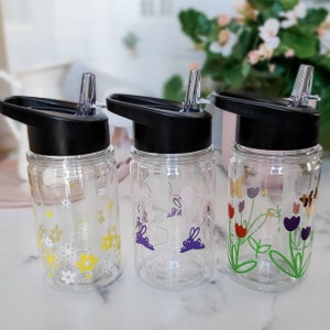 May include: Three clear plastic water bottles with black lids and built-in straws. Each bottle has a different floral design. One bottle has a floral design with yellow flowers, another has a floral design with purple flowers, and the third has a floral design with red and purple flowers.