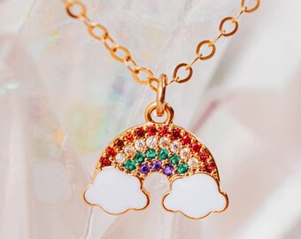 May include: A gold chain necklace with a rainbow charm. The rainbow is made of colorful gemstones and has white clouds on either side.