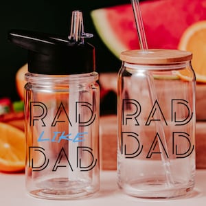 May include: Two clear glass water bottles with the text "RAD LIKE DAD" in black and blue. One bottle has a black lid and straw, the other has a bamboo lid and a clear straw. The background includes fruit.