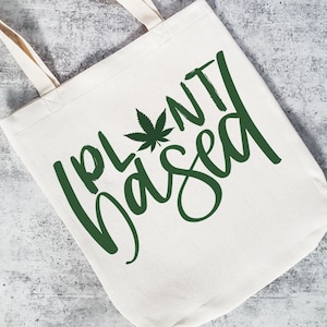 May include: A white canvas tote bag with a green marijuana leaf and the text "Plant Based" in green script.