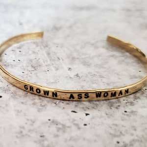 May include: A gold-toned bangle bracelet with the words "GROWN ASS WOMAN" engraved on the band.