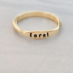 May include: A gold-colored ring with the word "feral" in black, stamped across the front. The ring has a smooth, polished finish and a simple, elegant design. The ring is a piece of jewelry.