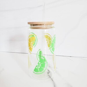 May include: A clear glass water bottle with a wooden lid and a straw. The bottle has a lemon and lime design.
