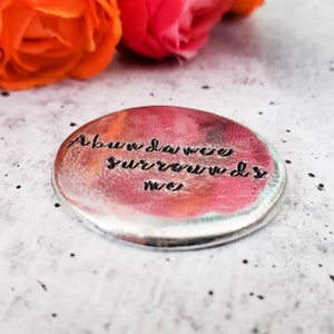 Peut inclure: Un charme en argent avec un fond aquarelle rose et orange. Le charme porte les mots "Above you were surrounded by" grav&eacute;s dessus.