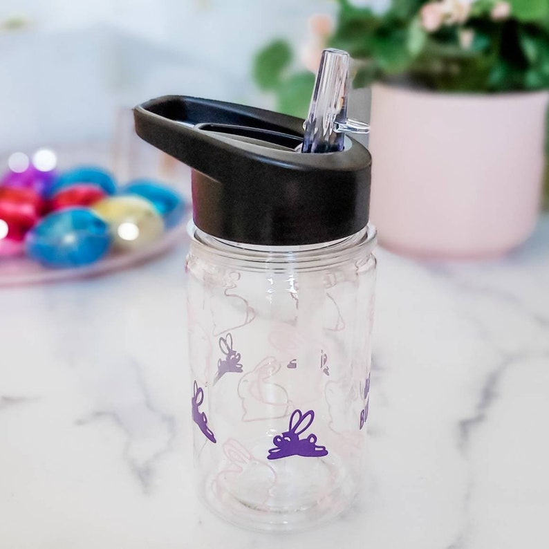 May include: A clear plastic water bottle with a black lid and a purple bunny design. The bottle has a built-in straw.