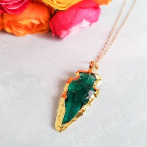 May include: A gold necklace with a green glass arrowhead pendant. The arrowhead is surrounded by a gold frame.