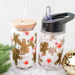 May include: Two clear glass tumblers with gingerbread man designs and red stars. One tumbler has a bamboo lid and a straw. The other tumbler has a black lid with a straw.
