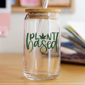 May include: A clear glass can with a bamboo lid and a green leaf design that says "Plant Based". A clear glass straw is in the can.