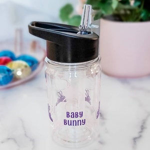 May include: Clear plastic water bottle with a black lid and a purple 'Baby Bunny' design. The bottle has a straw and a carrying handle.