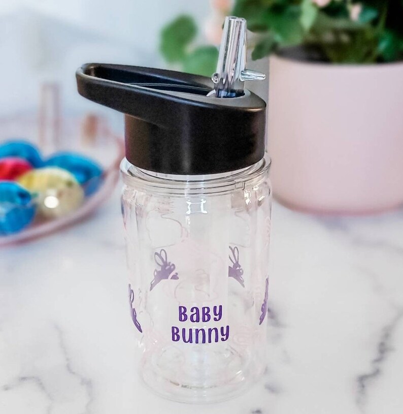 May include: Clear plastic water bottle with a black lid and a purple 'Baby Bunny' design. The bottle has a straw and a carrying handle.