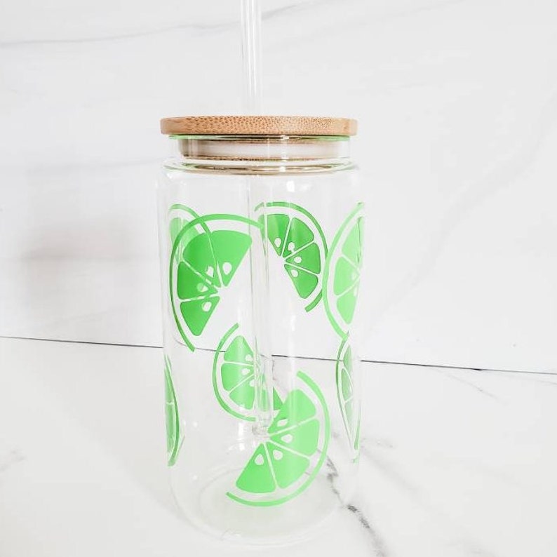 May include: A clear glass reusable water bottle with a bamboo lid and a green lime design. The bottle has a straw.