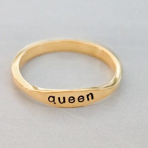 May include: A gold-colored ring with the word "queen" engraved on the band. The ring has a smooth, polished finish and a simple, elegant design. The ring is a piece of jewelry.