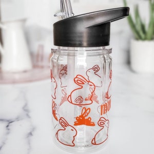 May include: A clear plastic water bottle with a black lid and a straw. The bottle has a pattern of orange bunnies on it.