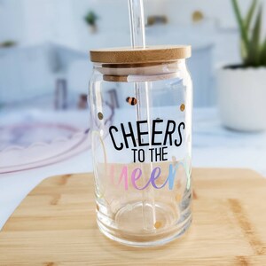 May include: A clear glass jar with a bamboo lid and a straw. The jar has gold glitter and the text "Cheers to the Queens" in rainbow colors.