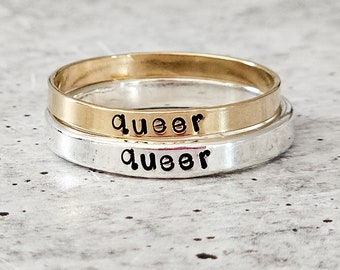 May include: Two stacked rings, one gold and one silver, with the word "queer" engraved on each.