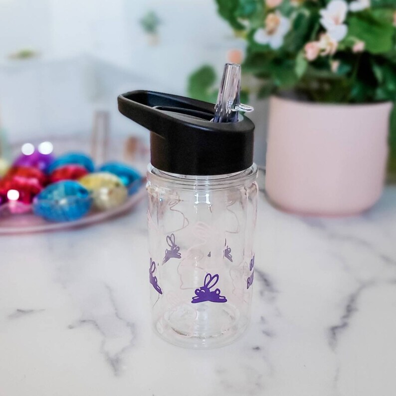 May include: A clear plastic water bottle with a black lid and a purple bunny design. The bottle has a straw and says "Happy Easter" on the side.