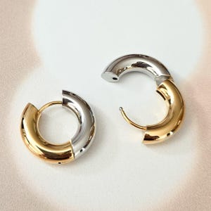 May include: Two-tone hoop earrings. One earring is closed, the other is open. The earrings are a combination of gold and silver. The earrings are on a white background.