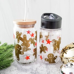 May include: Two clear glass tumblers with lids and straws. Both tumblers feature a gingerbread man design with red stars. One tumbler has a bamboo lid and the other has a black lid.