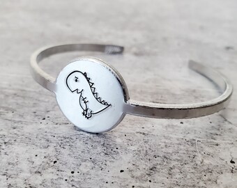 May include: Silver bangle bracelet with a round charm featuring a hand-drawn dinosaur design.
