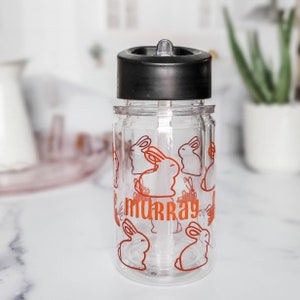 May include: A clear plastic water bottle with a black lid and a straw. The bottle has a red design of bunnies and the name "murray" printed on it.