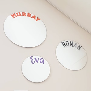 May include: Three round mirrors with names written on them. The top mirror has the name "MURRAY" written in orange letters. The middle mirror has the name "RONAN" written in black letters. The bottom mirror has the name "Eva" written in purple letters.