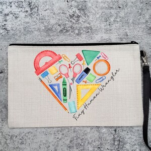 May include: A beige canvas zippered pouch with a black zipper and a black strap. The pouch has a heart-shaped design filled with colorful school supplies, including a protractor, scissors, crayons, pencils, and a ruler. The text "Tiny Human Wrangler" is written in black cursive below the heart.