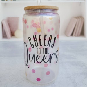 May include: A clear glass tumbler with a bamboo lid and a straw. The tumbler has a pink and gold confetti pattern and the text "Cheers to the Queers".