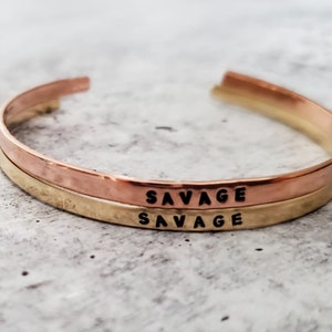 May include: Two stacked gold and rose gold bracelets with the word "SAVAGE" engraved on each.
