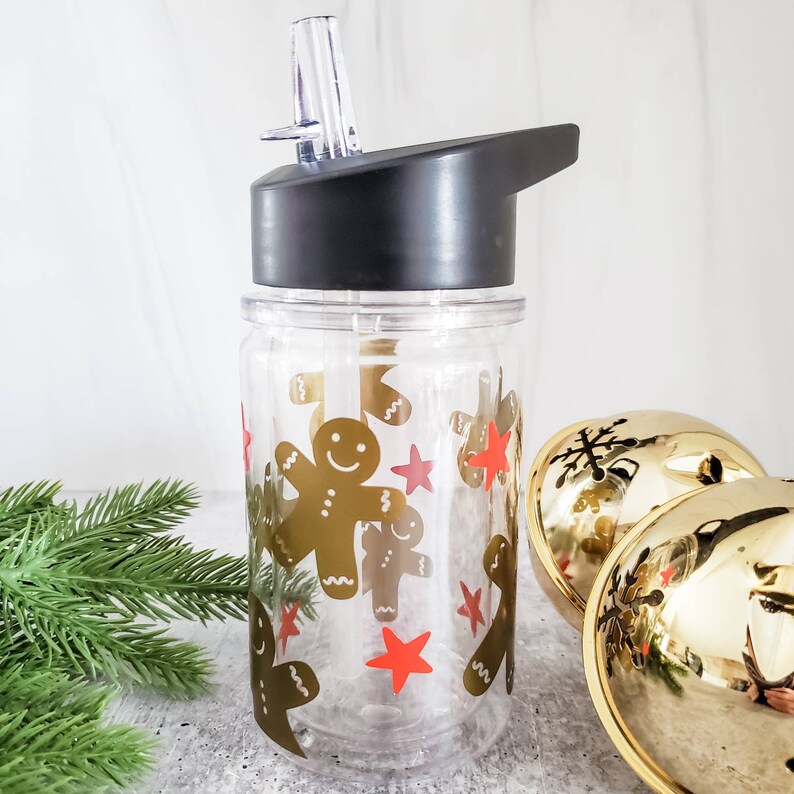 May include: A clear plastic water bottle with a black lid and a straw. The bottle is decorated with gingerbread men and red stars.