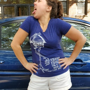 Pode incluir: Uma mulher vestindo uma camiseta azul com um gráfico branco de uma casa em pernas de frango. O gráfico inclui o texto "Избушка Избушка" e "Стань, мы тебе передом в лесу заглянем". Ela está vestindo shorts brancos e tem esmalte de unha azul.