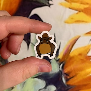 May include: A sticker featuring a cartoon frog wearing a brown cowboy hat. The frog is brown with a white outline. The sticker is held between two fingers.