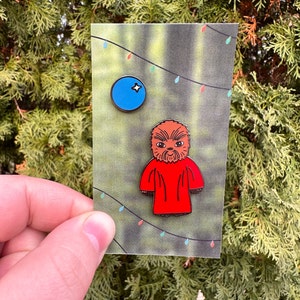 Puede incluir: Dos pines de esmalte sobre una tarjeta. Un pin es un círculo azul con una estrella blanca. El otro pin es un personaje de dibujos animados con una túnica roja y pelaje marrón. La tarjeta tiene un fondo verde con luces de cadena.
