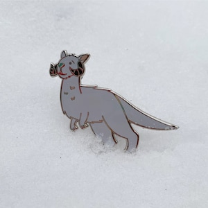 以下が含まれることがあります： 曲がった角、長い尾、赤い口を持つ様式化された動物のライトグレーのエナメルピン。 ピンは銀色の輪郭で、雪の背景に設定されています。