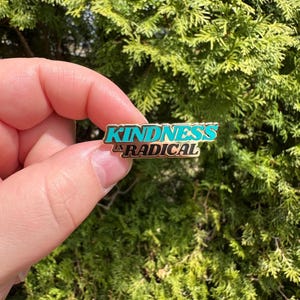 Op de afbeelding: Een rechthoekige emaille pin met de woorden "KINDNESS IS RADICAL" in turquoise en gouden letters. De pin wordt tegen een achtergrond van groen gebladerte gehouden.
