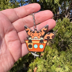 May include: A metal keychain featuring a cartoon character with orange and brown fur, peeking out of an orange container. The container has a black circle, two light blue circles, and a silver spoon. The keychain has a silver chain and ring.
