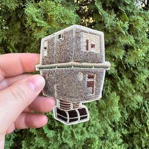 May include: Embroidered patch featuring a brown, boxy, robot-like design with window details. The patch has a textured appearance and is outlined with a green border. The bottom section has a series of brown and black rectangles.