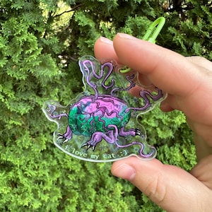 Op de afbeelding: Een heldere acryl sleutelhanger met een grillig, buitenaards ontwerp in paars, groen en roze. De sleutelhanger heeft een groene sluiting en de tekst "i love my cat :)". De achtergrond is een wazig beeld van groen gebladerte.