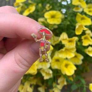 May include: A gold-colored metal pin featuring a stylized robot character with a red head and torso. The robot has a light pink body and is holding up one hand. The pin is set against a background of yellow flowers.