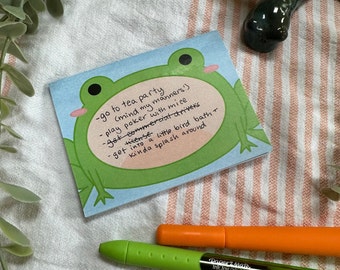 May include: A square notepad featuring a cartoon frog design with the text "-go to tea party (mind my manners!) - play poker with mice - get commercial drivers license - get into a little bird bath + kinda splash around". Two pens are also visible.