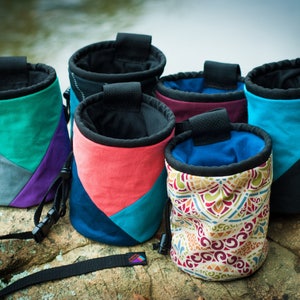 May include: Six colorful climbing chalk bags with different patterns and designs. The bags are made of fabric and have a drawstring closure. They are sitting on a rock surface.