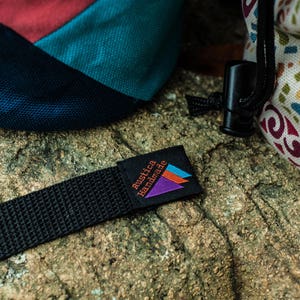May include: A black strap with a small black label that says "Rustica Handmade" in orange and white text. The label has a purple, blue, and orange triangle design.