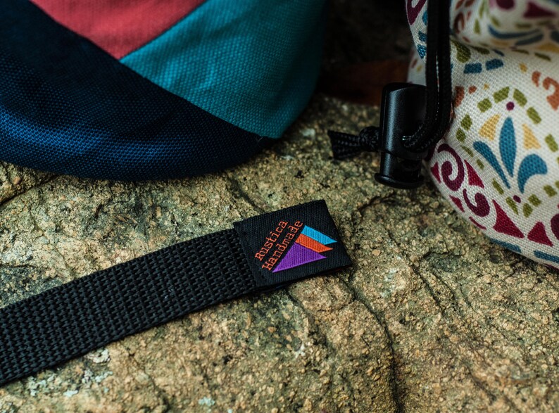 May include: A black strap with a small black label that says "Rustica Handmade" in orange and white text. The label has a purple, blue, and orange triangle design.