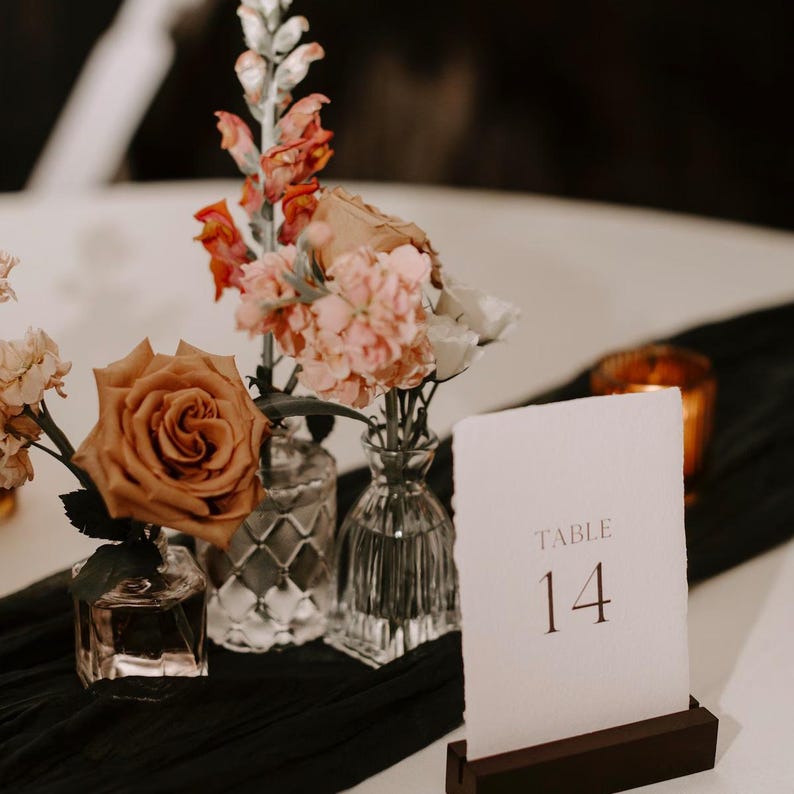 May include: A table number card with the number 14 printed in brown on a white background. The card is sitting on a wooden stand. The card is surrounded by a floral arrangement of pink, orange, and brown flowers in small glass vases.