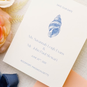 May include: Cream-colored wedding invitation with blue text and a seashell illustration. The text reads "Save Our Date," the names of the couple, the wedding date, and the location, Rockport, Massachusetts.