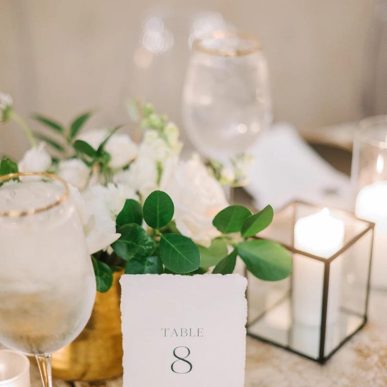 May include: A white table number card with gold trim and the text "TABLE 8" on a white background. The card is sitting on a gold vase with white flowers and green leaves. There is a clear glass of water with a gold rim and a candle in a glass lantern in the background.