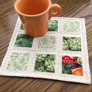 May include: A white quilted coaster with a green and white floral pattern. The coaster has a stitched border and is decorated with squares of different floral patterns. An orange mug is sitting on top of the coaster.