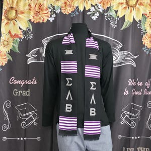 May include: Black graduation stole with purple and white stripes and Greek letters. The stole is draped over a black long-sleeved shirt. The background is a dark curtain with floral and graduation-themed graphics.