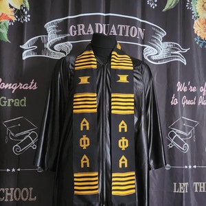 May include: Black graduation gown with a gold and black graduation stole with the letters "AΦA" on it. The background is a black chalkboard with the words "Graduation", "Congrats Grad", "School", "We're off to Great Places", and "Let the" written on it.