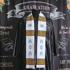 May include: A white and gold graduation stole with the letters "AΦA" in black and white. The stole has a black background with gold stripes.